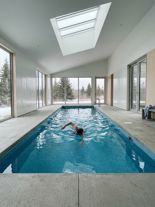 A person swimming in a long indoor lap pool inside a modern home featuring floor-to-ceiling windows overlooking a snowy forest and a large skylight.