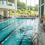 Swimmer in modern indoor lap pool with lane dividers, featuring floor-to-ceiling windows overlooking green landscape, exposed wooden ceiling beams, natural light, pool ladder, and multi-level facility with white architectural design