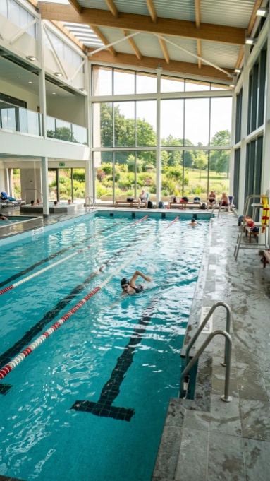 Swimmer in modern indoor lap pool with lane dividers, featuring floor-to-ceiling windows overlooking green landscape, exposed wooden ceiling beams, natural light, pool ladder, and multi-level facility with white architectural design
