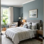 Bright bedroom with soft blue walls, white bedding, blue patterned area rug, wooden nightstand, dark dresser, and armchair by window